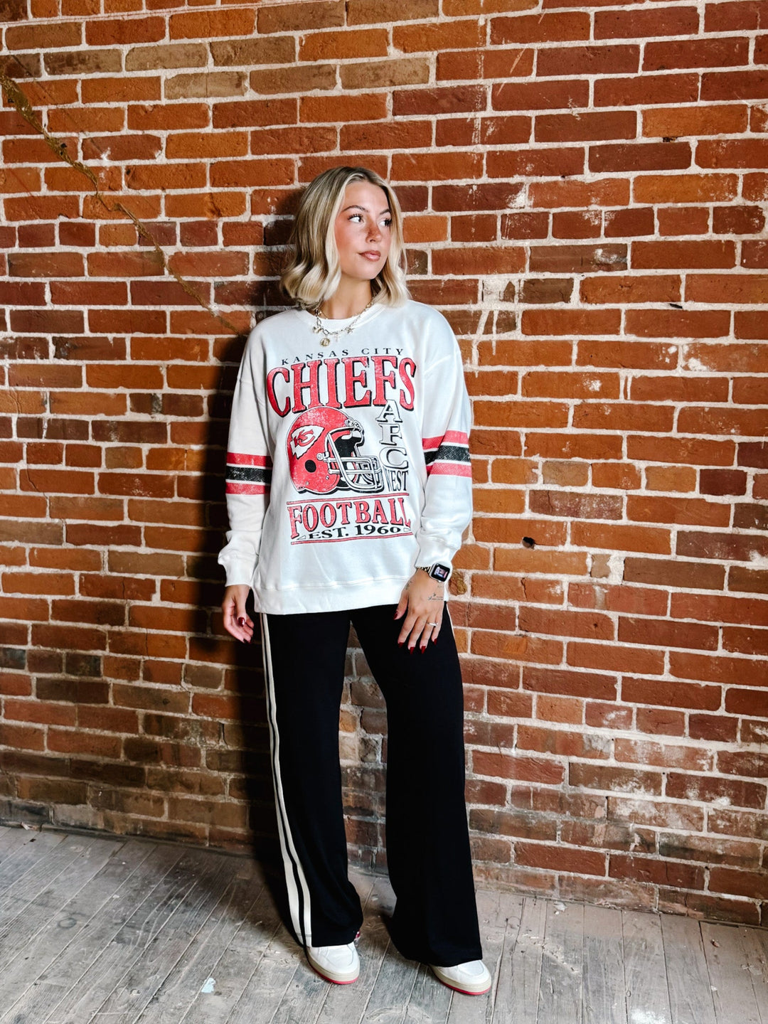 A woman stands against a red brick wall wearing a white long-sleeve graphic sweatshirt with red and navy lettering that reads &
