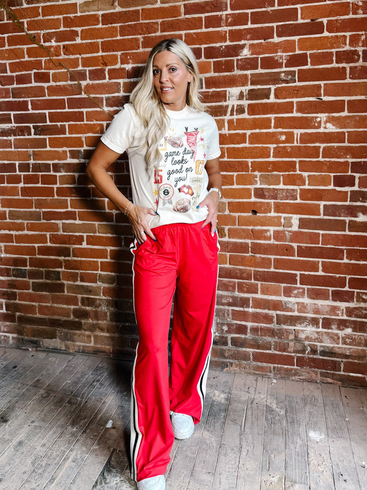 A woman posing in front of a red brick wall wearing a white graphic t-shirt and bright red track pants with white side stripes.