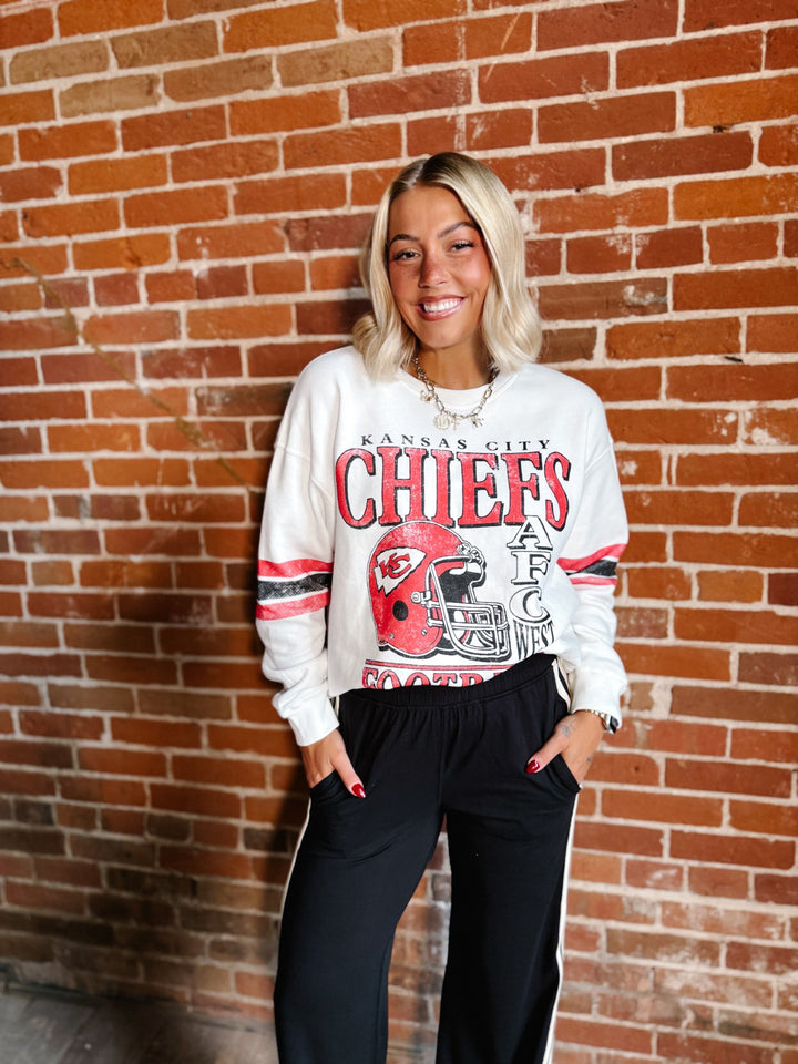 A woman stands against a red brick wall wearing a white Kansas City Chiefs sweatshirt with a red and black graphic print, paired with black sweatpants with white side stripes.
