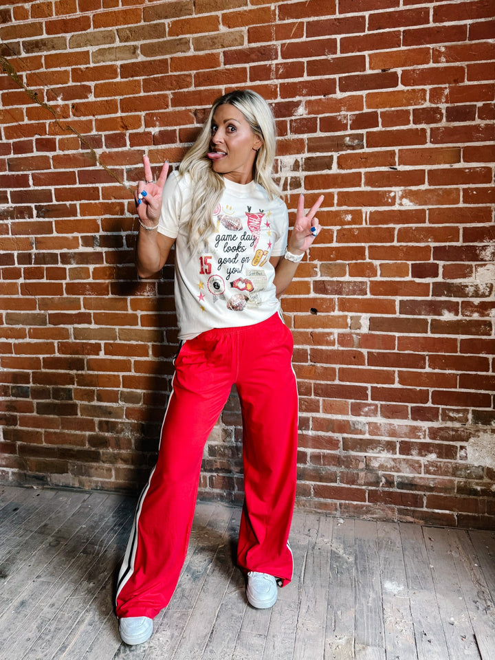 A woman posing in front of a brick wall wearing a cream graphic t-shirt and bright red track pants with white side stripes.