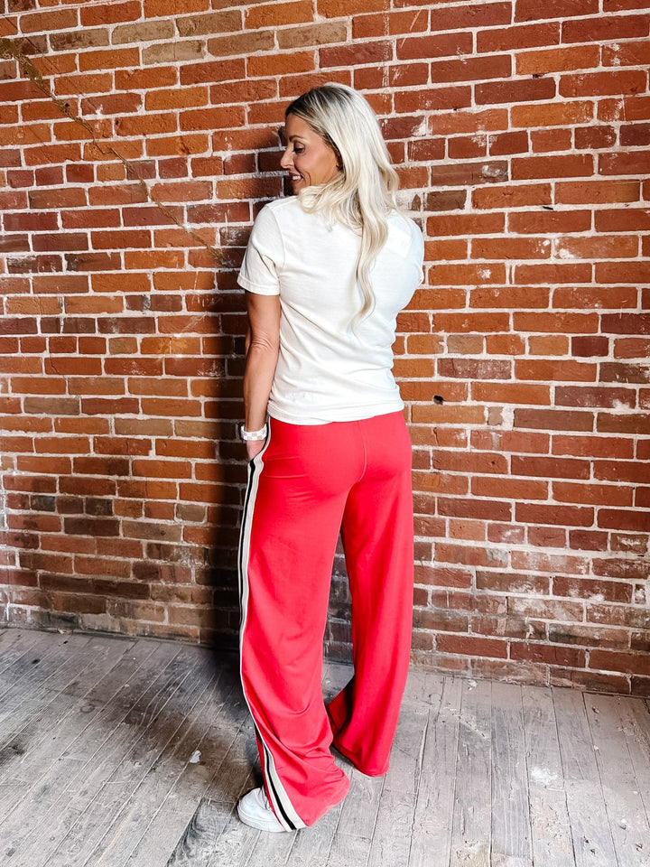A woman with blonde hair stands with her back to the camera, wearing bright red wide-leg sweatpants with white side stripes, a white t-shirt, and white sneakers, posing against a red-brown brick wall.