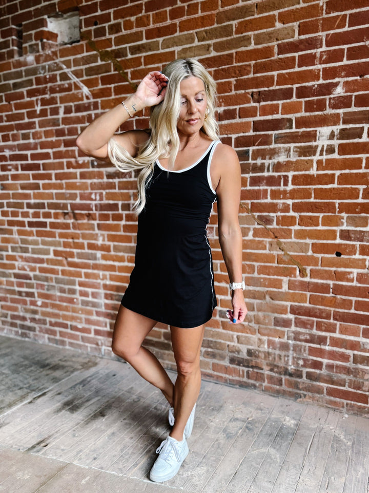 A woman wearing a black skortsie dress with a scoop neckline and contrast trim, stylishly posing indoors.