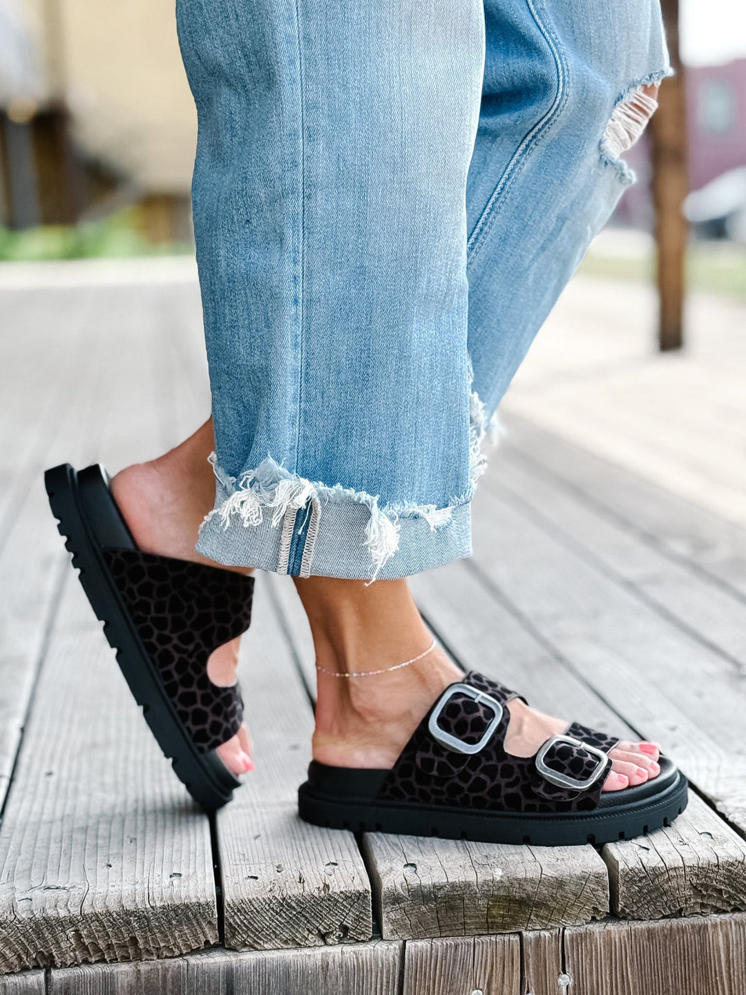 A person wearing light blue ripped jeans and black leopard-print slide sandals with silver buckle straps, standing on a weathered wooden deck.