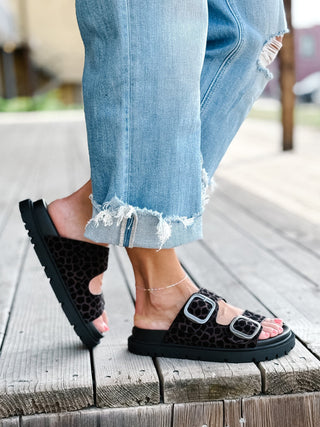 A person wearing light blue ripped jeans and black leopard-print slide sandals with silver buckle straps, standing on a weathered wooden deck.