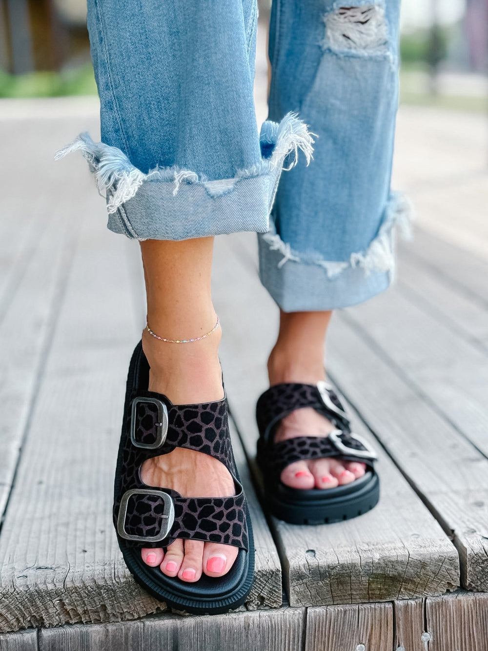 A person wearing black platform sandals with a leopard print pattern and double buckles, paired with distressed light blue jeans, standing on a wooden deck.