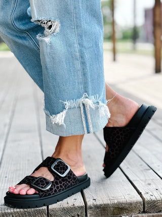 A person wearing black platform slide sandals with a brown leopard print pattern and silver buckle straps, paired with light blue ripped jeans, standing on a wooden deck.