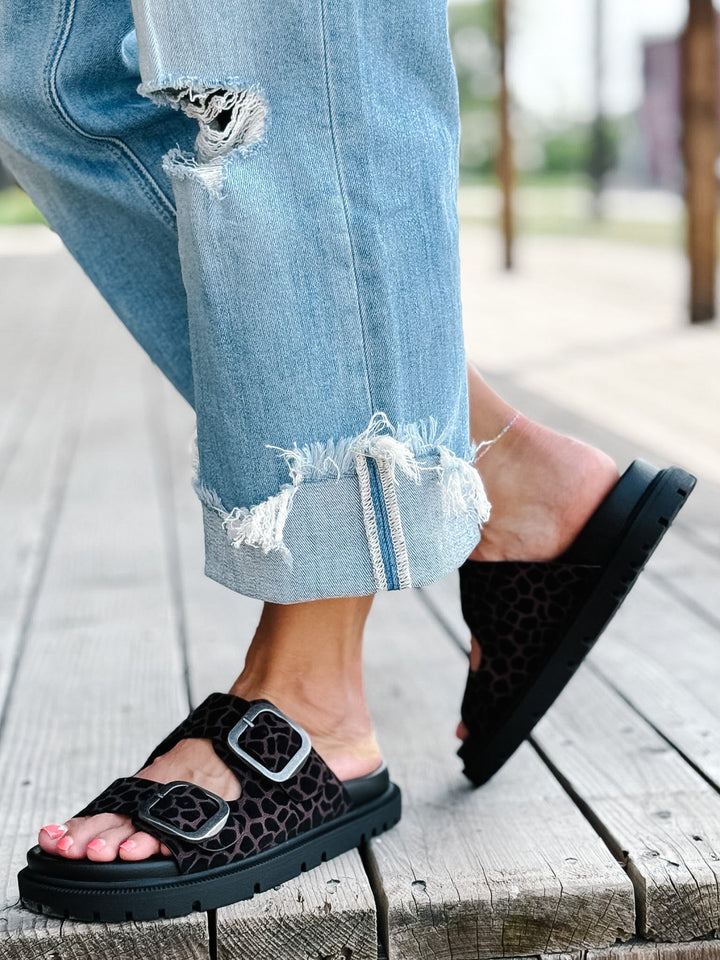 A person wearing black platform slide sandals with a brown leopard print pattern and silver buckle straps, paired with light blue ripped jeans, standing on a wooden deck.