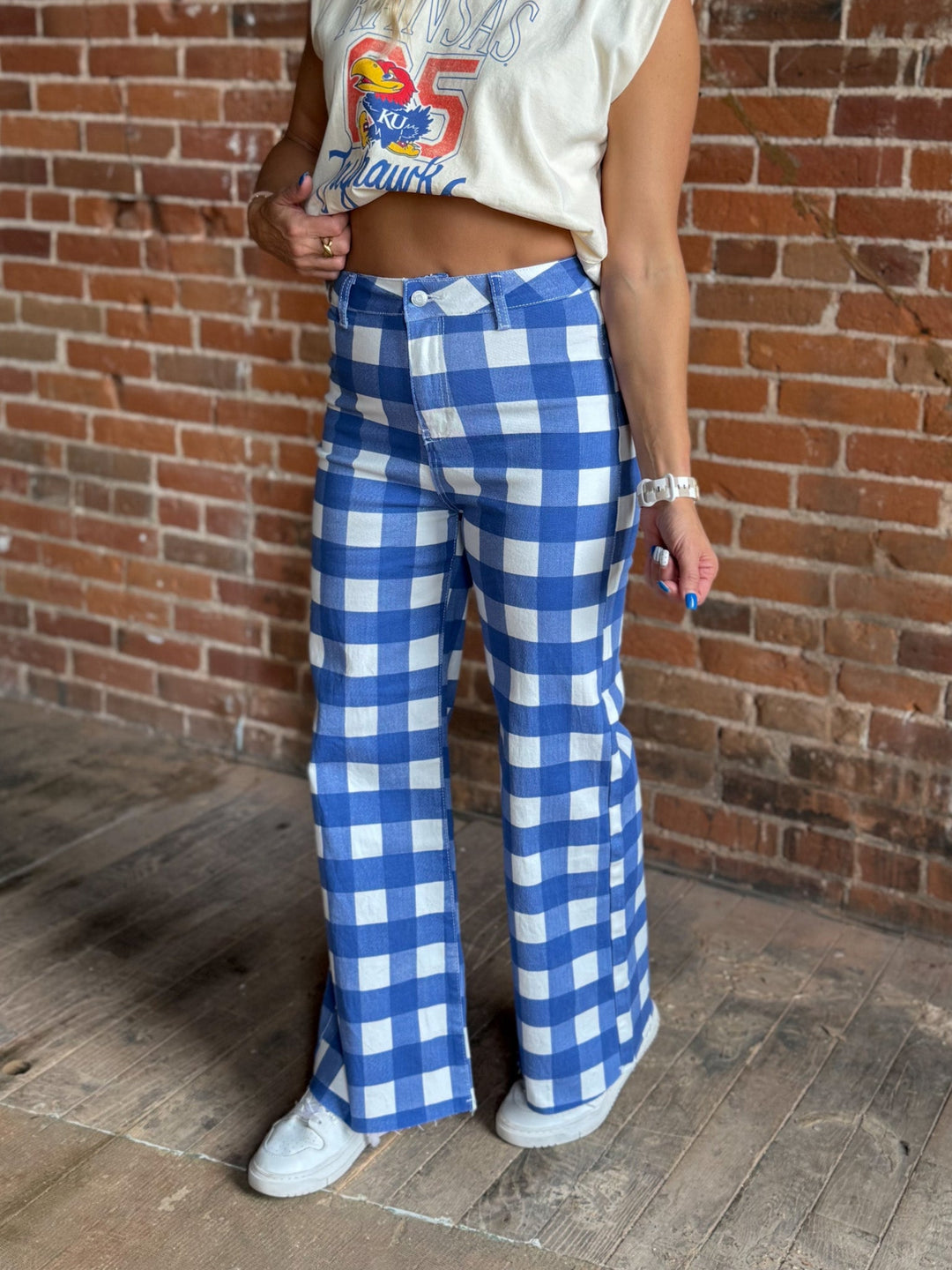 A person wearing blue and white gingham wide-leg pants, a cream sleeveless crop top, and white sneakers, standing against a brick wall.