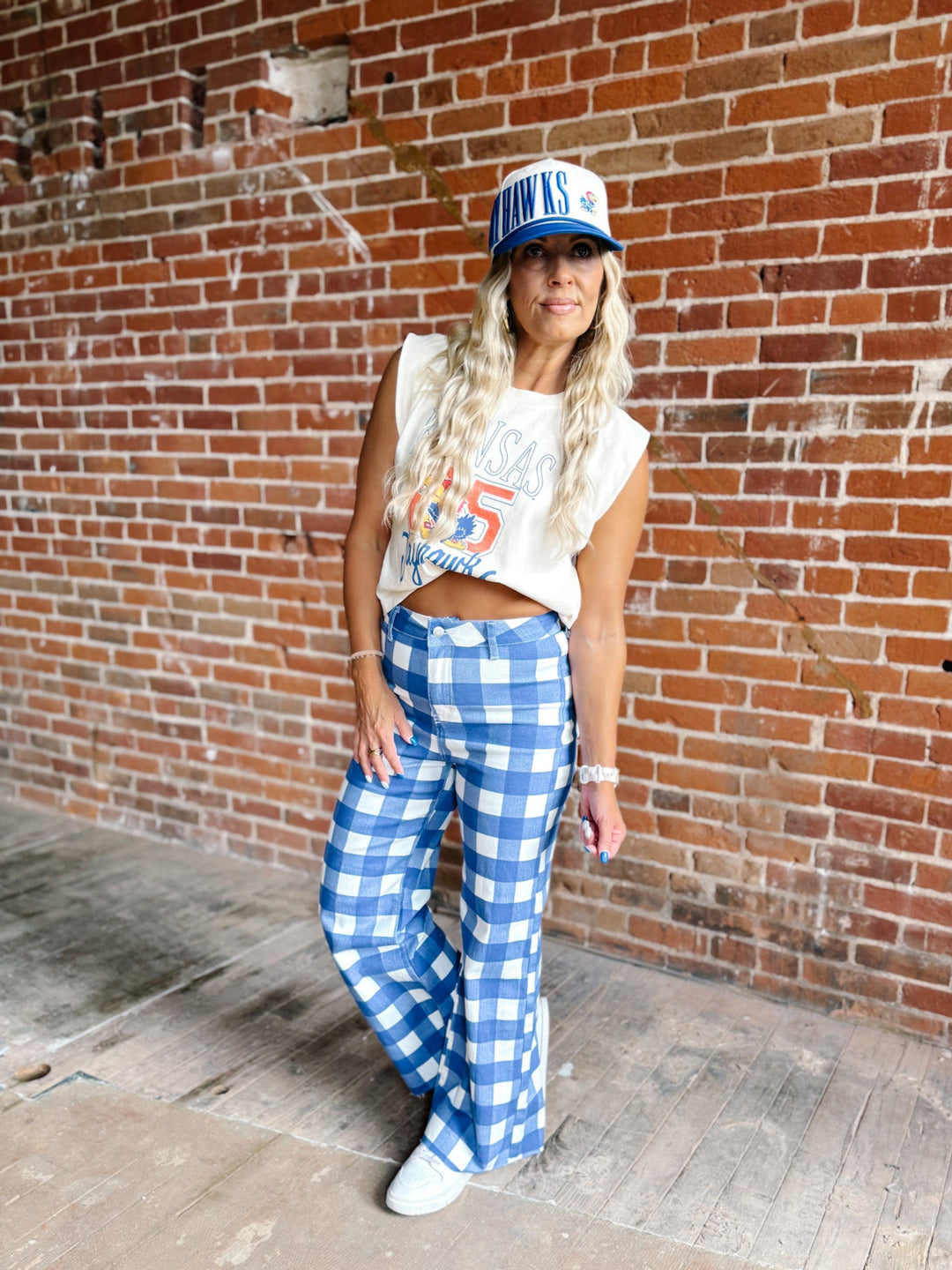 A woman stands against a red brick wall wearing a white cropped tank top with a blue and red graphic, blue and white gingham wide-leg pants, white sneakers, and a blue and white trucker hat.