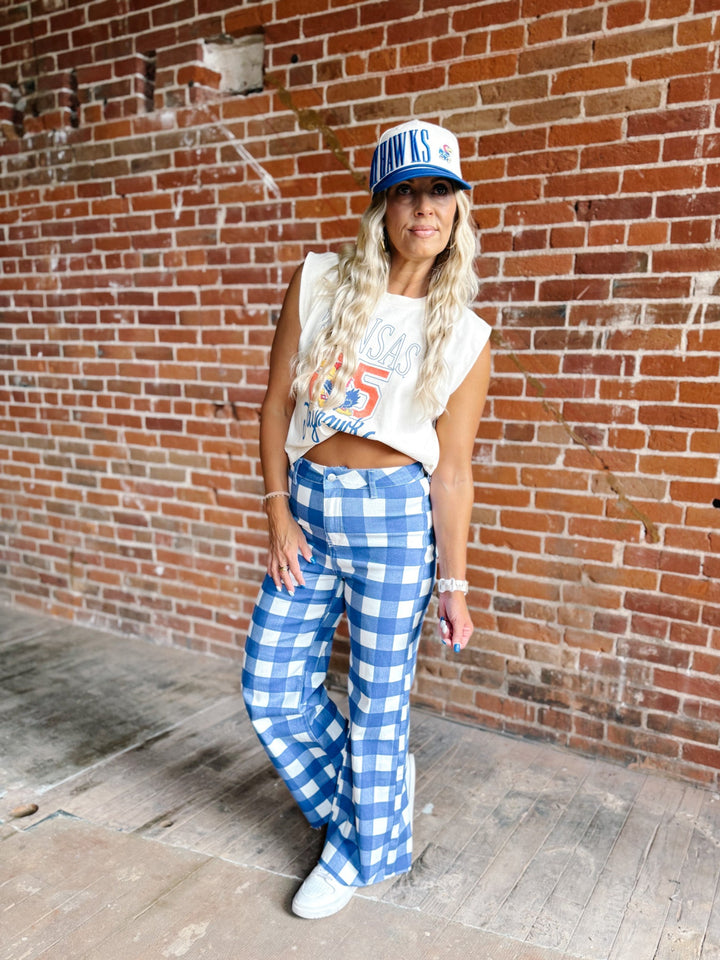 A woman stands against a red brick wall wearing a white cropped tank top with a blue and red graphic, blue and white gingham wide-leg pants, white sneakers, and a blue and white trucker hat.