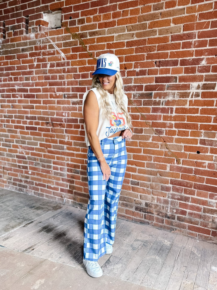 A woman stands against a red brick wall wearing a white and blue gingham checkered pair of wide-leg pants, a white graphic tank top, a blue and white baseball cap, and white sneakers.