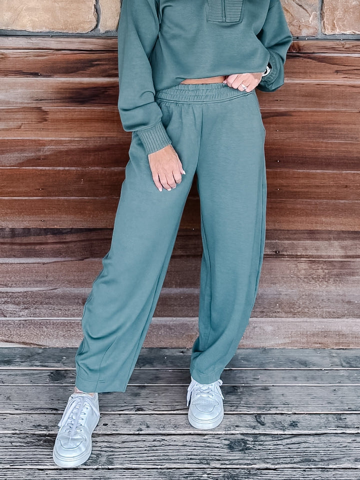 A person wearing a matching sage green sweatshirt and jogger pants set, paired with white sneakers, standing against a rustic wooden wall.