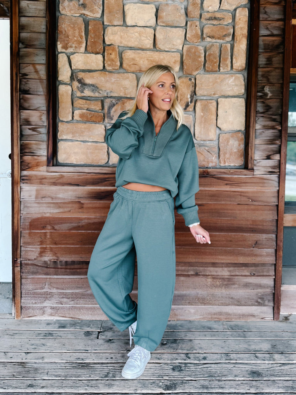 A person wearing a matching sage green sweatshirt and jogger pants set, paired with white sneakers, standing against a rustic wooden wall.