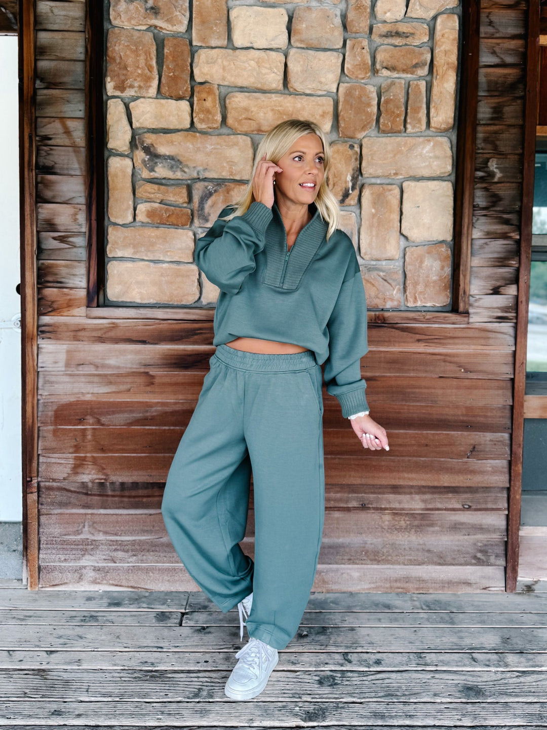 A person wearing a matching sage green sweatshirt and jogger pants set, paired with white sneakers, standing against a rustic wooden wall.