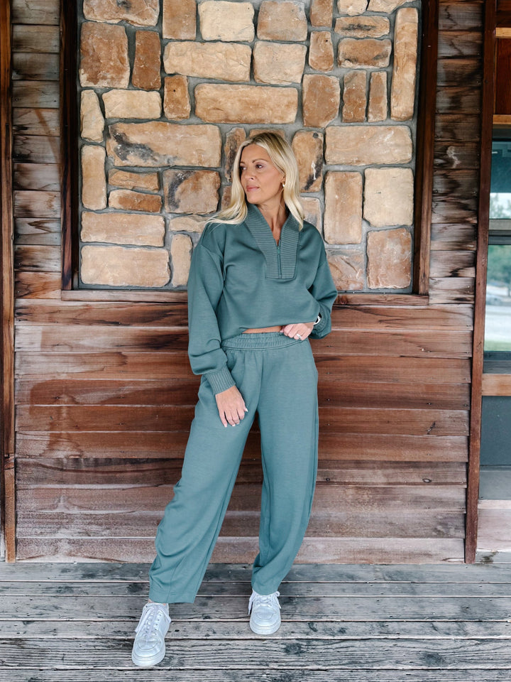 A person wearing a matching sage green sweatshirt and jogger pants set, paired with white sneakers, standing against a rustic wooden wall.