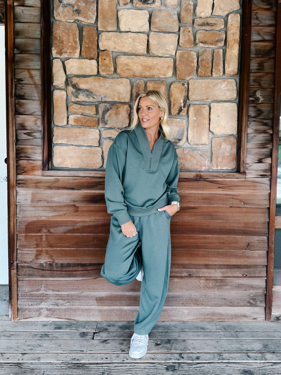 A person wearing a matching sage green sweatshirt and jogger pants set, paired with white sneakers, standing against a rustic wooden wall.