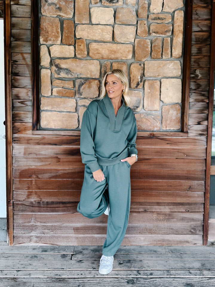 A person wearing a matching sage green sweatshirt and jogger pants set, paired with white sneakers, standing against a rustic wooden wall.