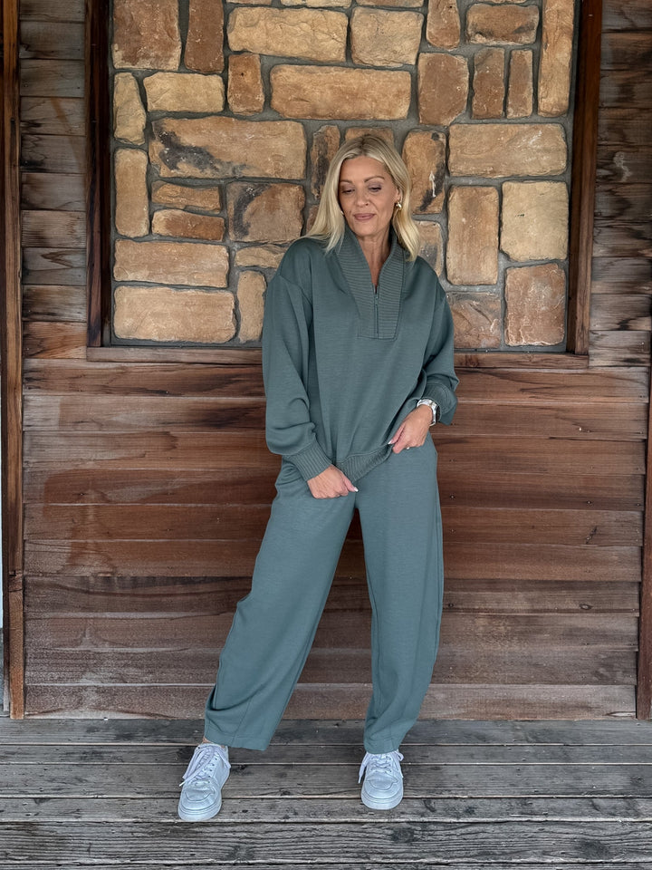 A person wearing a matching sage green sweatshirt and jogger pants set, paired with white sneakers, standing against a rustic wooden wall.