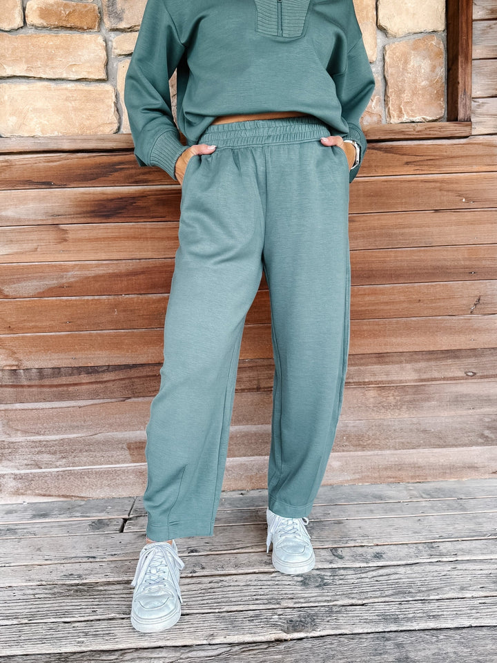 A person wearing a matching sage green sweatshirt and jogger pants set, paired with white sneakers, standing against a rustic wooden wall.