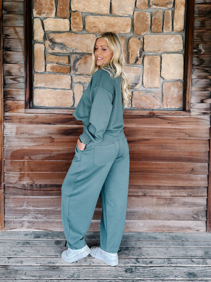 A woman modeling a sage green two-piece lounge set consisting of a long-sleeve top and matching wide-leg pants, standing on a wooden deck in front of a rustic stone and wood wall.