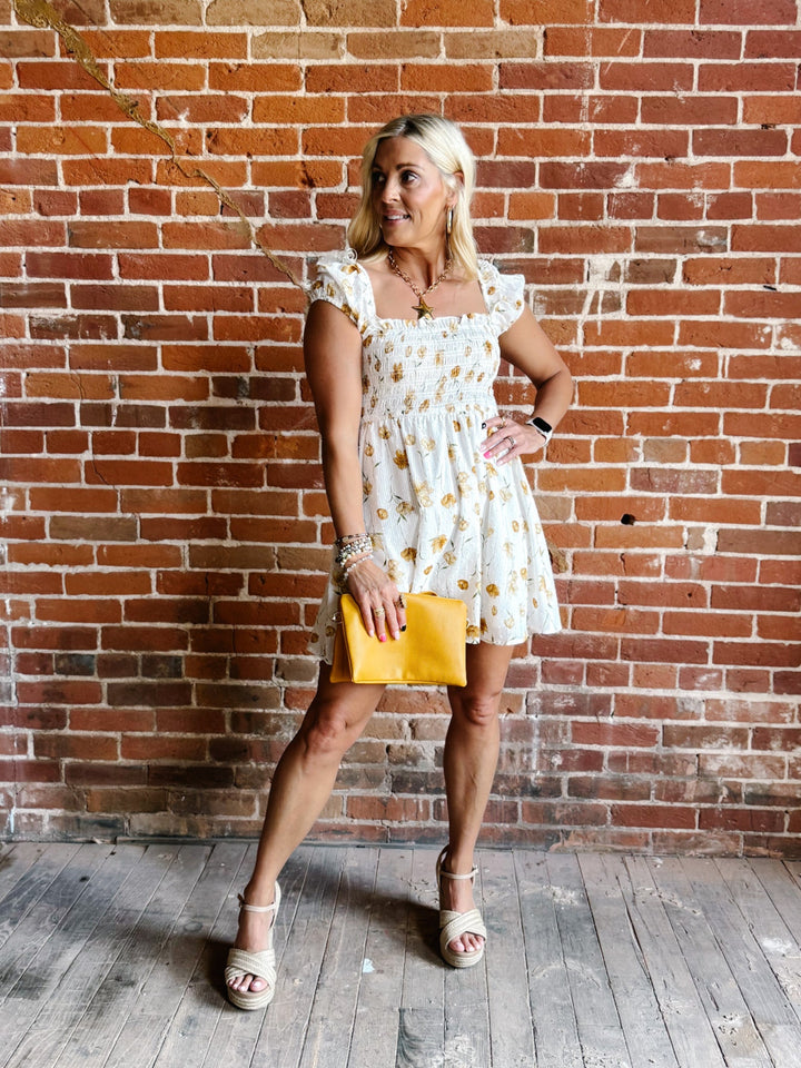 A woman wearing a white mini dress with a floral print and square neckline, standing against a brick wall.