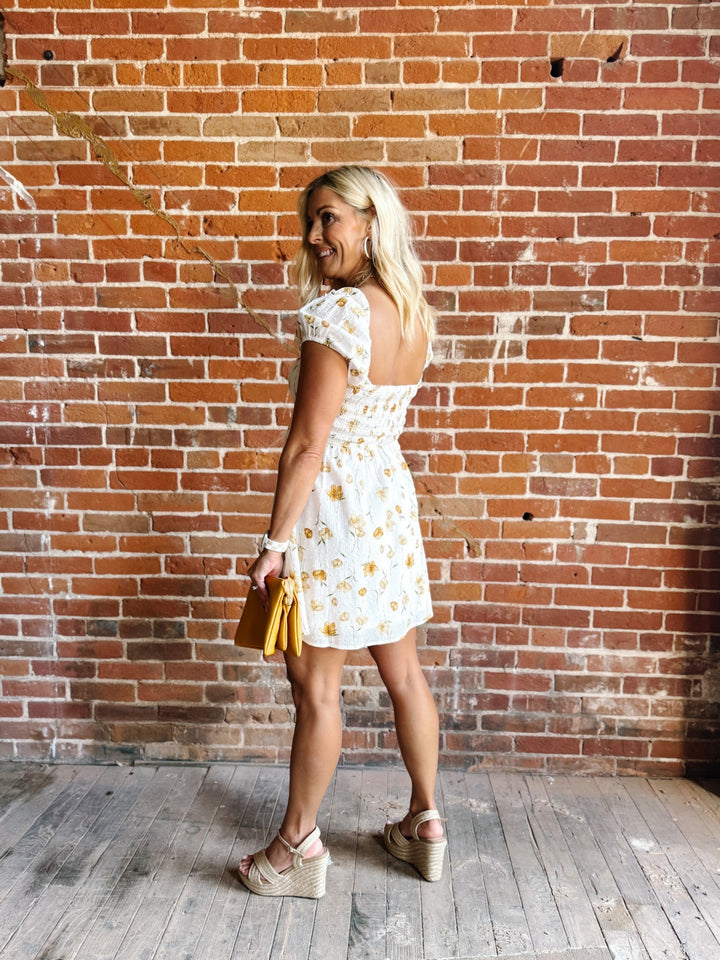 A woman wearing a white mini dress with a floral print and square neckline, standing against a brick wall.