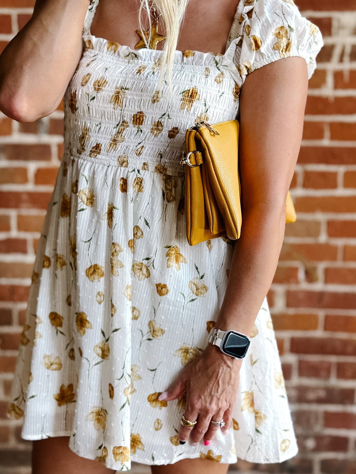 A woman wearing a white mini dress with a floral print and square neckline, standing against a brick wall.