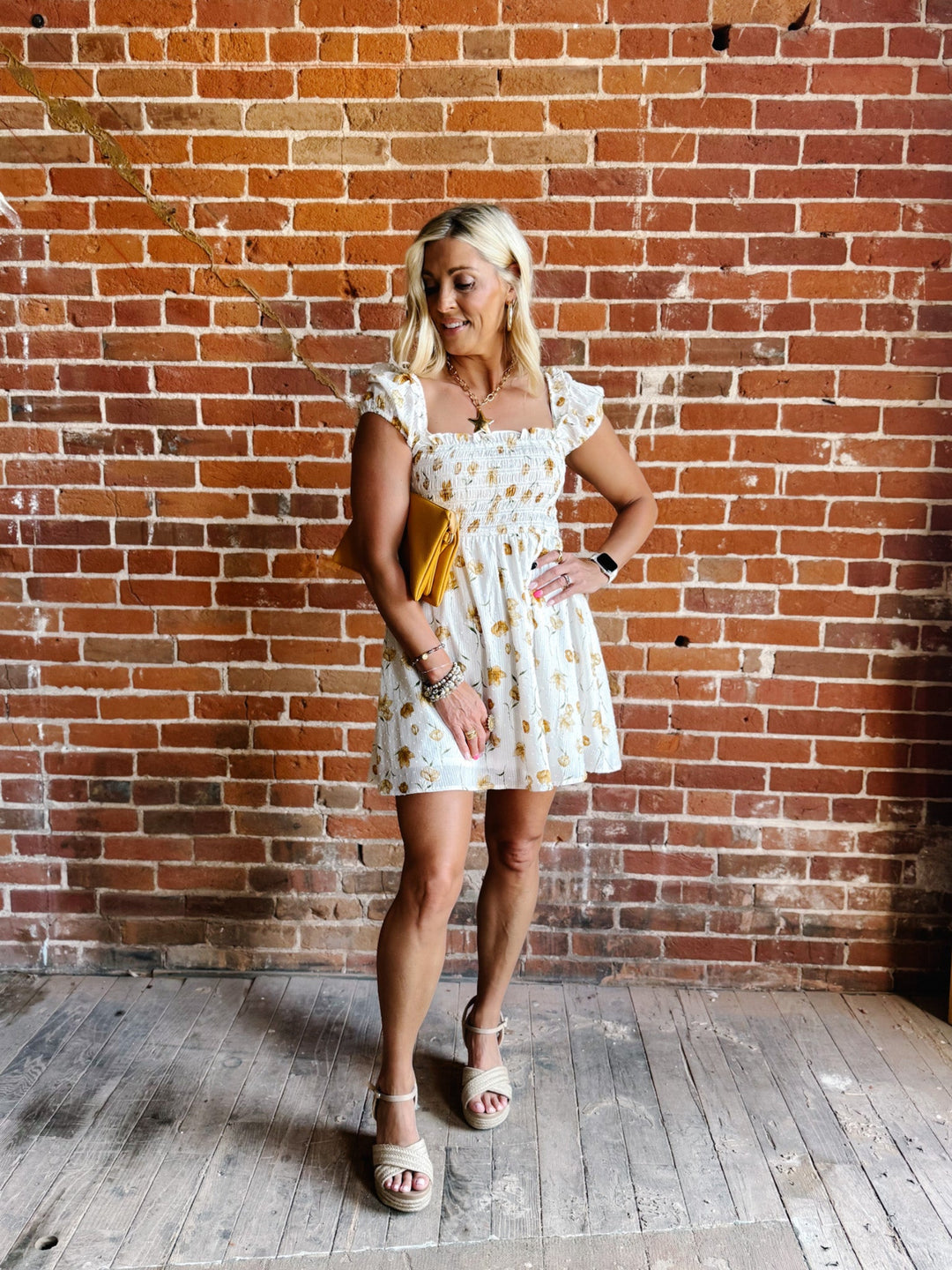 A woman wearing a white mini dress with a floral print and square neckline, standing against a brick wall.