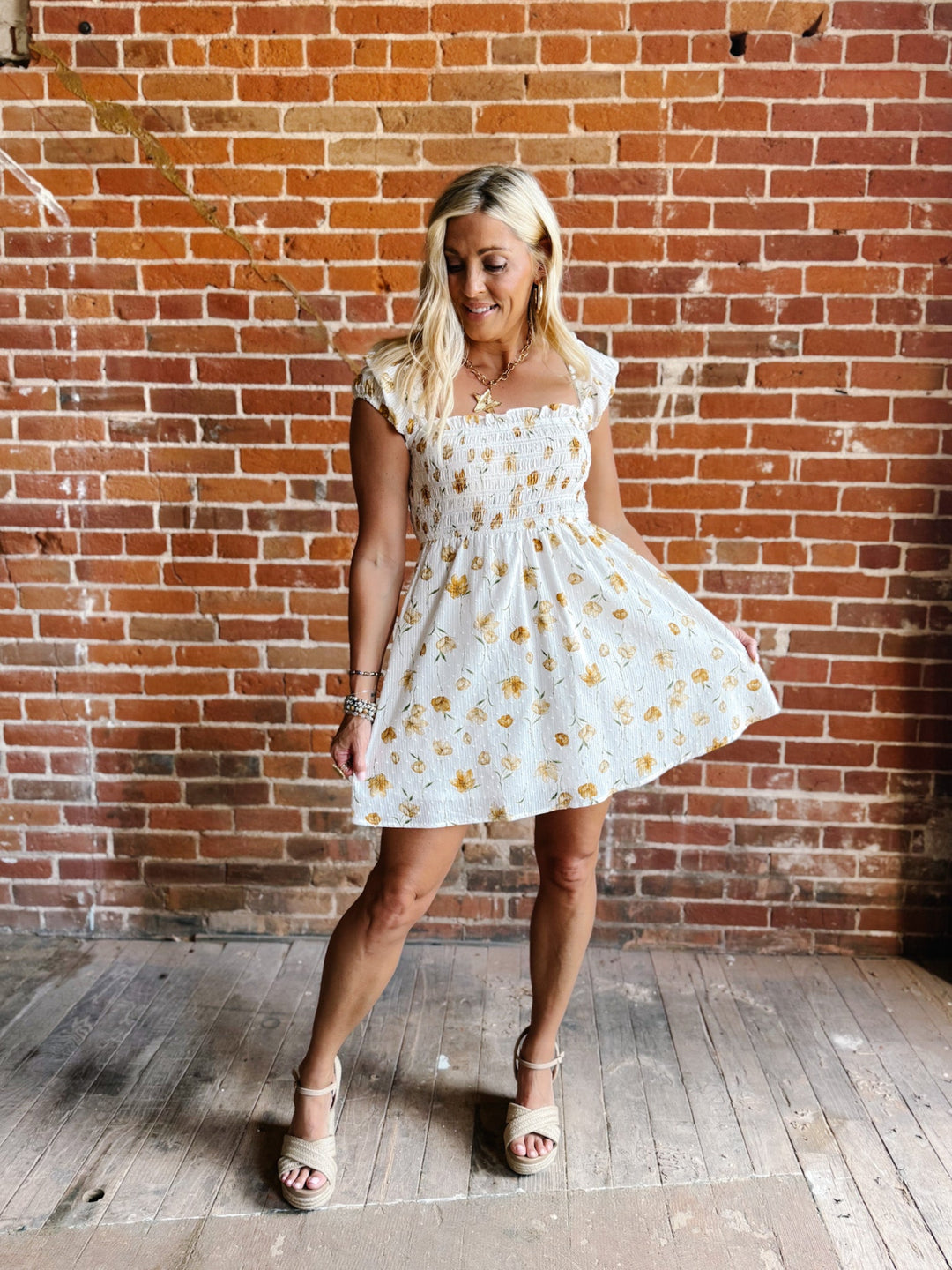 A woman wearing a white mini dress with a floral print and square neckline, standing against a brick wall.