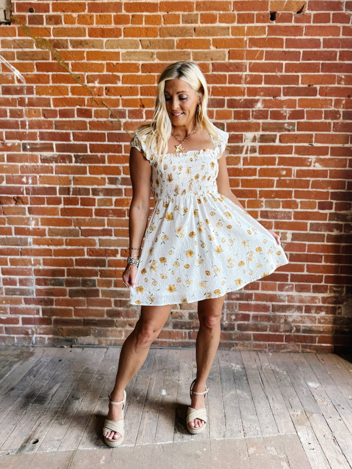 A woman wearing a white mini dress with a floral print and square neckline, standing against a brick wall.