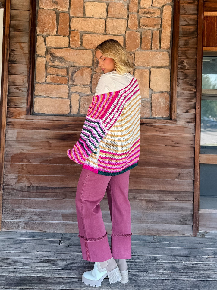 A woman stands on a wooden porch in front of a stone wall, wearing a chunky knit striped cardigan with bright pink, orange, yellow, white, and blue stripes, paired with dusty rose pants and white platform shoes.