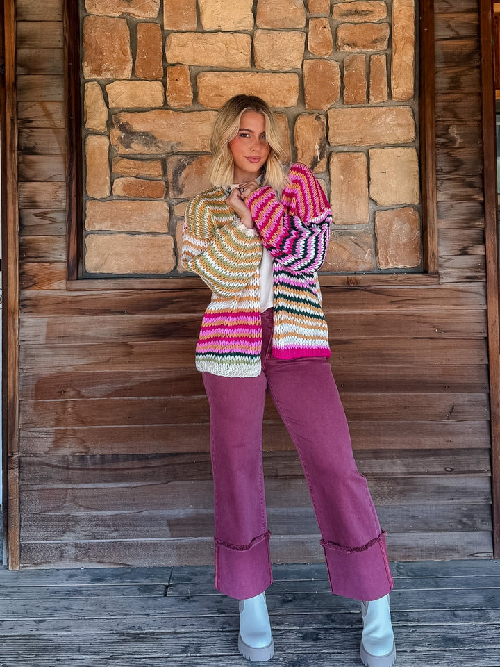 A woman stands on a rustic wooden porch in front of a stone wall, wearing a colorful striped knit cardigan and mauve wide-leg pants with white boots.