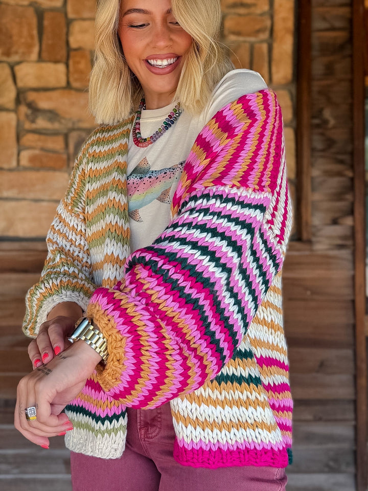 A woman wearing a chunky knit cardigan with bold zigzag stripes in pink, white, black, green, and tan, layered over a graphic t-shirt and mauve pants, standing in front of a rustic wooden door.
