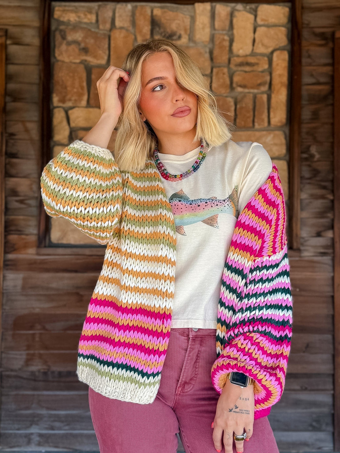 A woman wearing a chunky, multicolor striped knit cardigan with one sleeve in pink, teal, and white, and the other in beige, orange, and white, paired with a white graphic t-shirt and dusty rose jeans, standing in front of a rustic wooden and stone wall.