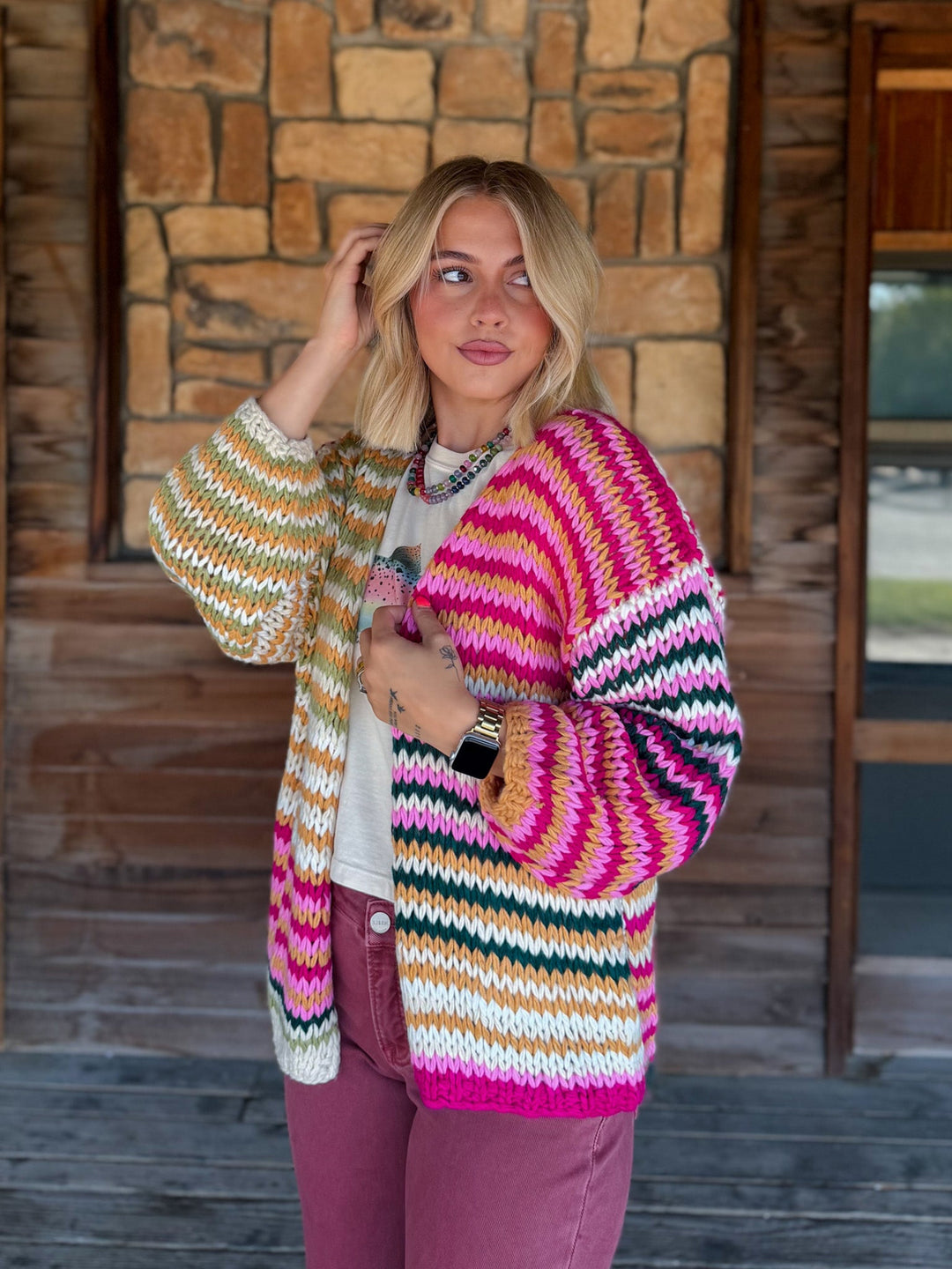 A woman stands on a rustic wooden porch wearing a chunky hand-knit cardigan with vibrant horizontal stripes in pink, orange, yellow, green, and white, paired with mauve pants.