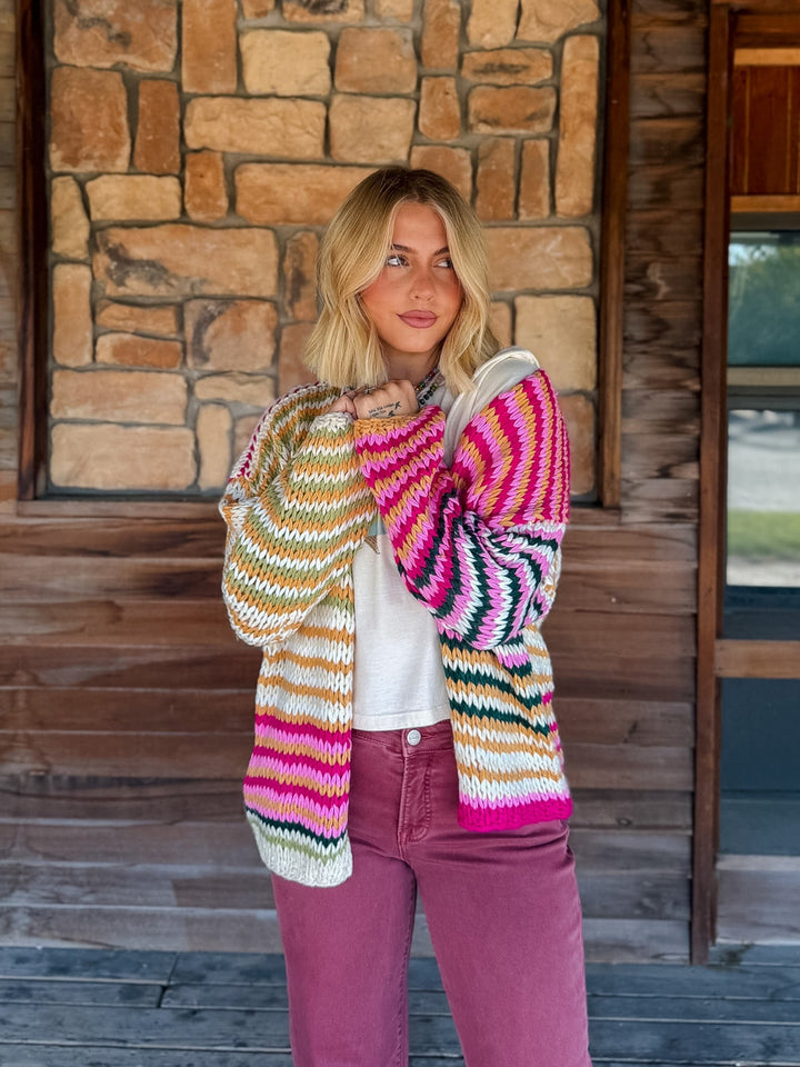 A woman wearing a vibrant, multicolored striped knit cardigan over a white t-shirt and mauve pants, standing in front of a rustic stone and wood wall.