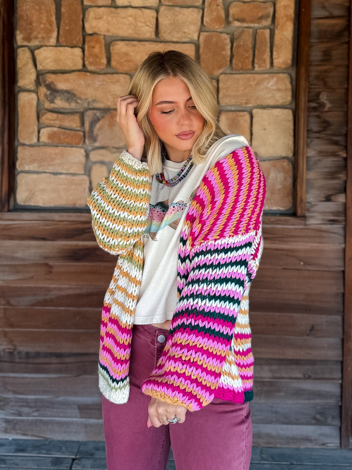A woman wearing a vibrant, multicolor striped knit cardigan with pink, yellow, green, and white stripes, layered over a white graphic t-shirt and paired with mauve pants, standing in front of a rustic stone and wood wall.