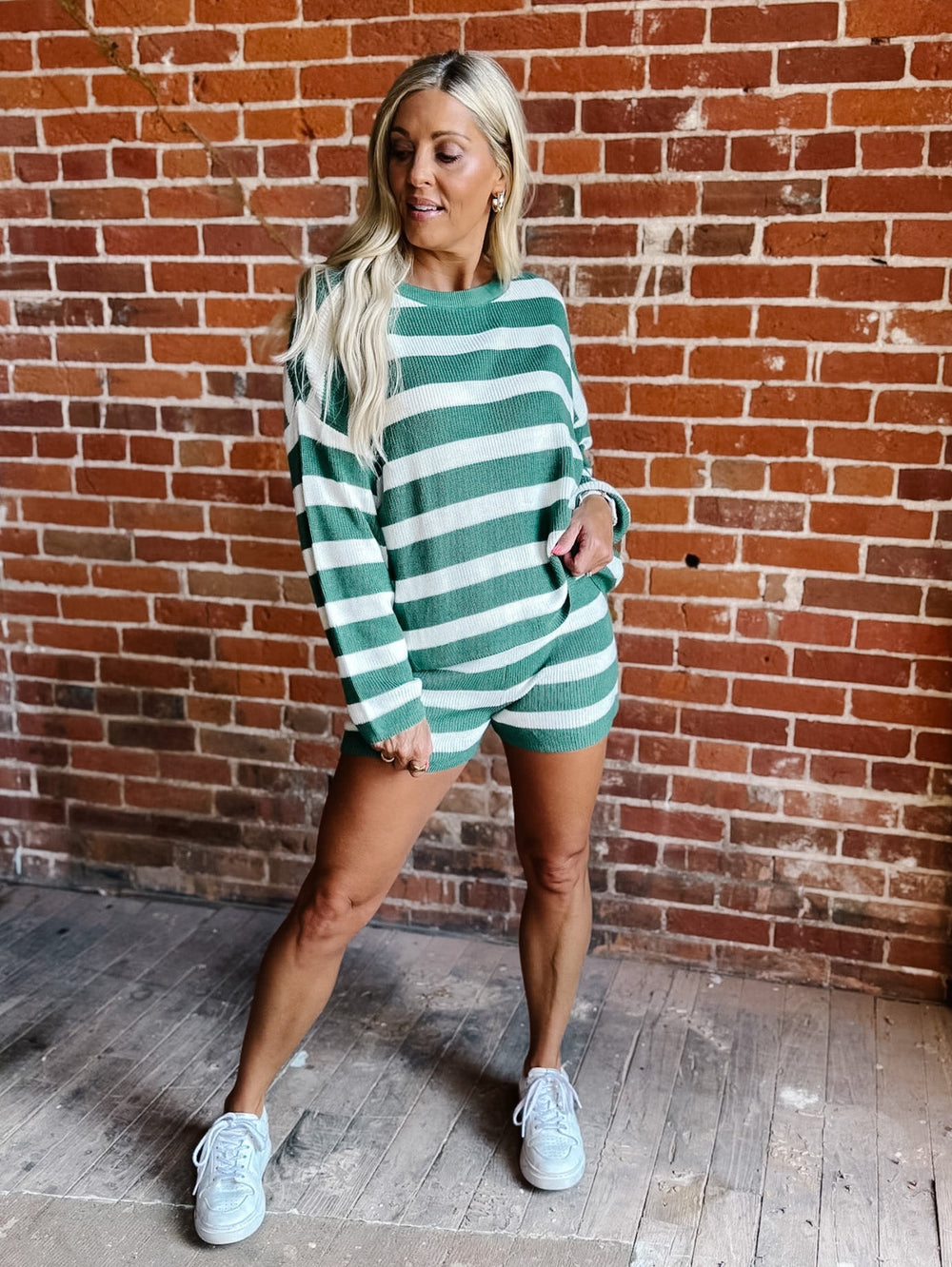 A woman modeling a green and white horizontally striped knit romper with long sleeves, standing in front of a red brick wall.