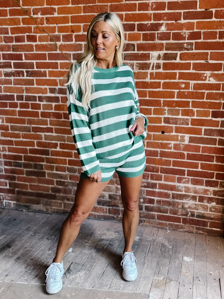 A woman modeling a green and white horizontally striped knit romper with long sleeves, standing in front of a red brick wall.