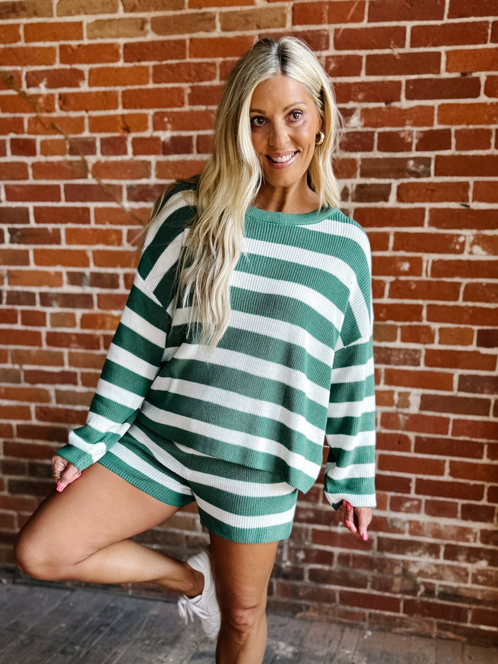 A woman with blonde hair wearing an oversized green and white striped knit sweater dress, posing in front of a red brick wall.