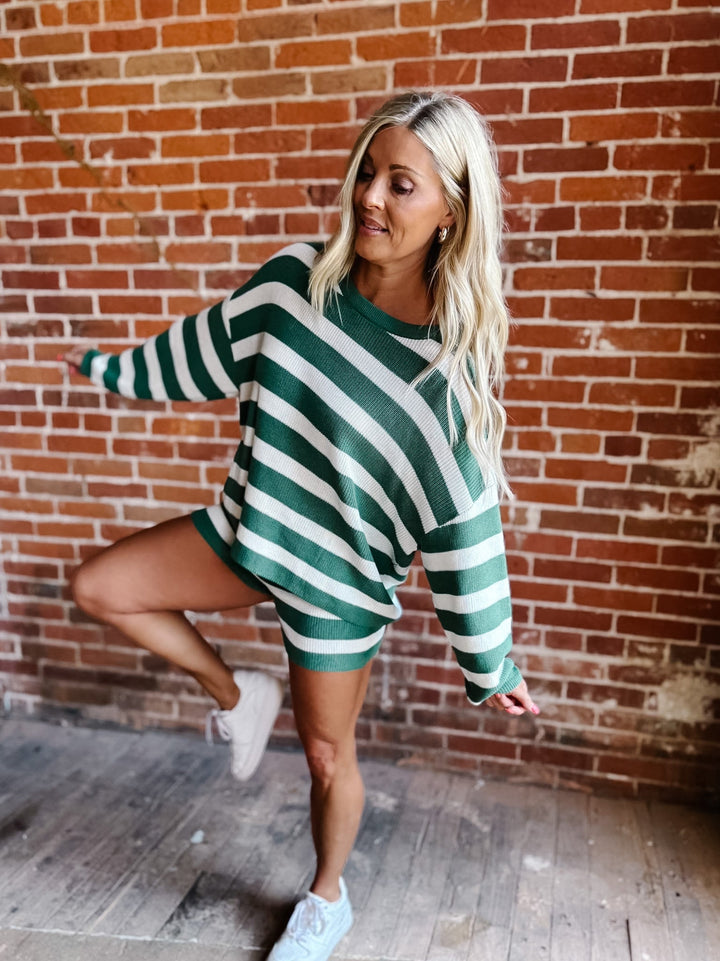 A woman posing in front of a brick wall wearing a green and white striped long-sleeve mini dress with white sneakers.