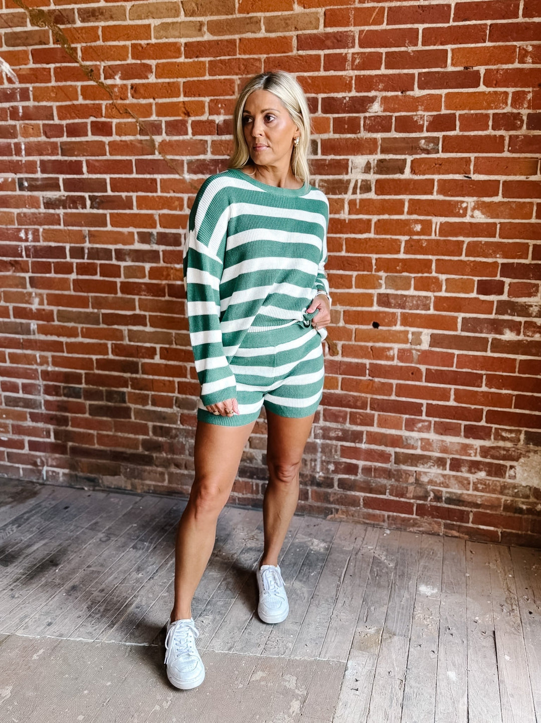 A woman modeling a green and white striped long-sleeve romper with matching shorts, standing against a red brick wall background.
