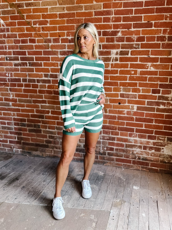 A woman modeling a green and white striped long-sleeve romper with matching shorts, standing against a red brick wall background.