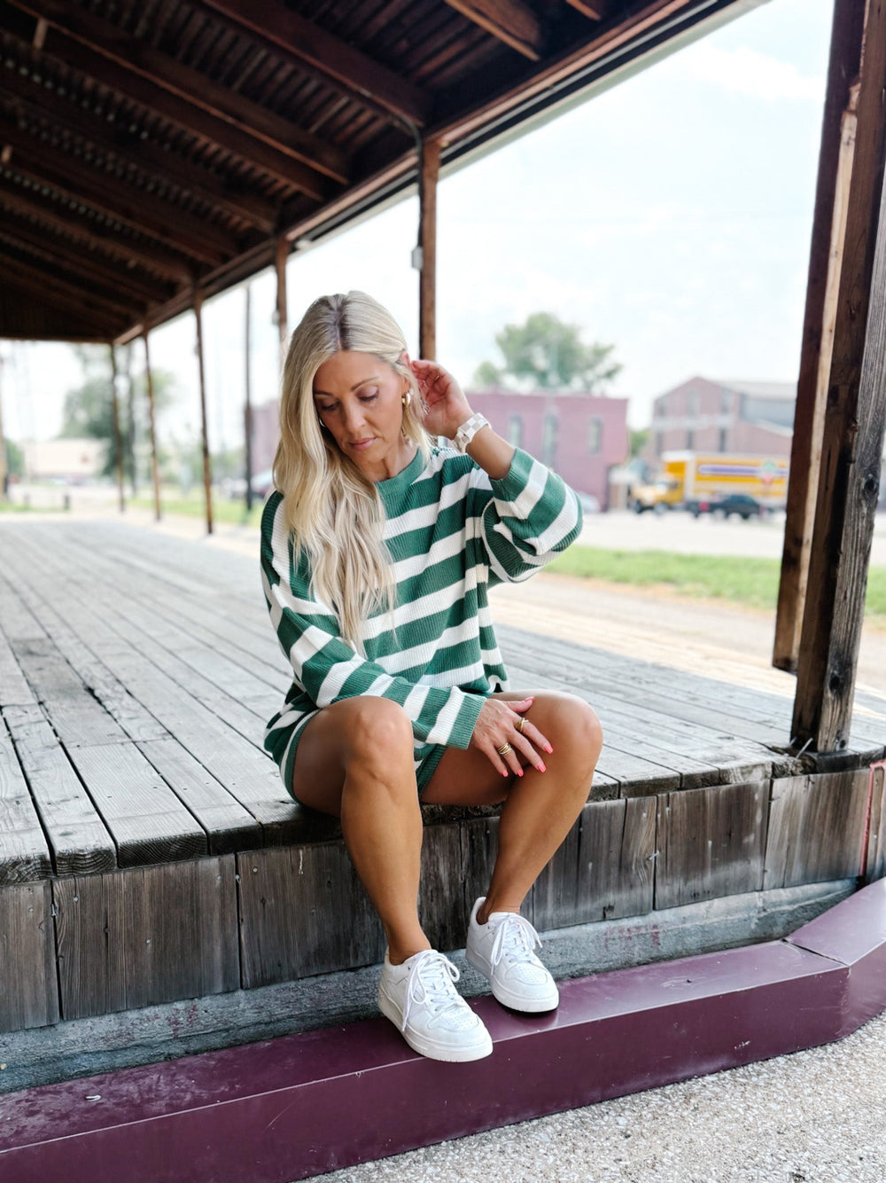 A woman with blonde hair sits on a weathered wooden platform under a covered structure, wearing a green and white horizontally striped long-sleeve tunic top and white sneakers.