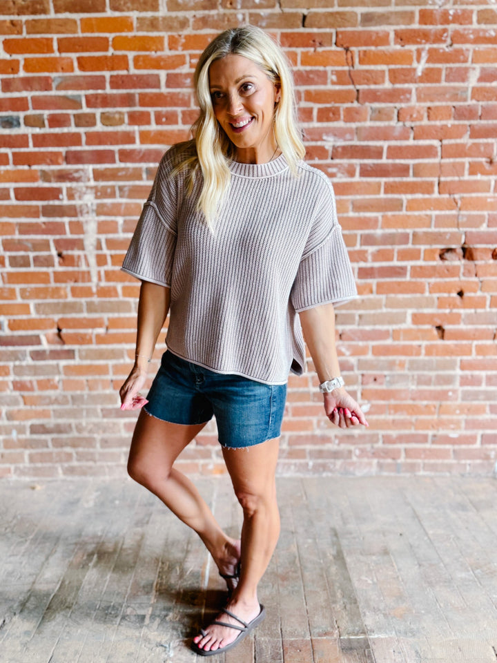 Woman wearing a soft knit sweater with exposed seams and short sleeves, standing against a brick wall.