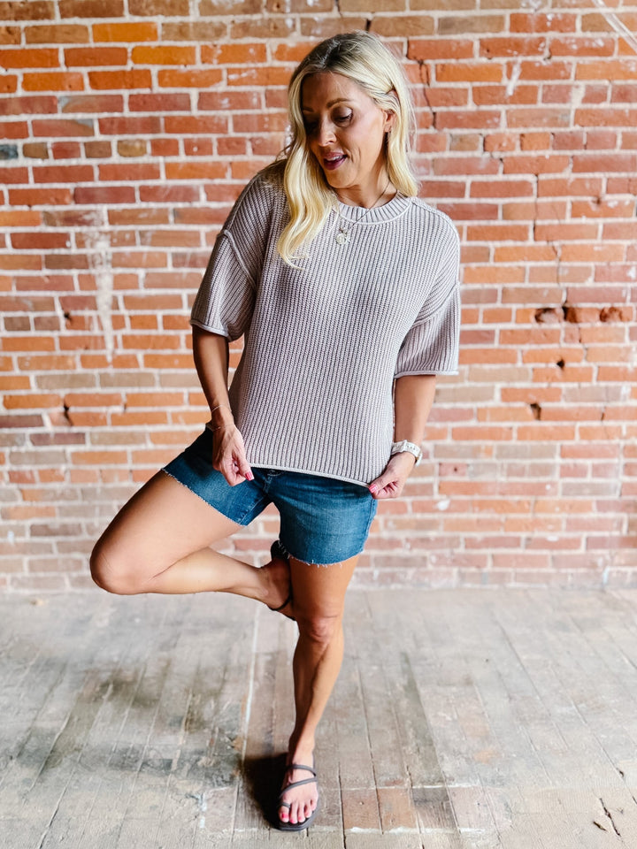 Woman wearing a soft knit sweater with exposed seams and short sleeves, standing against a brick wall.