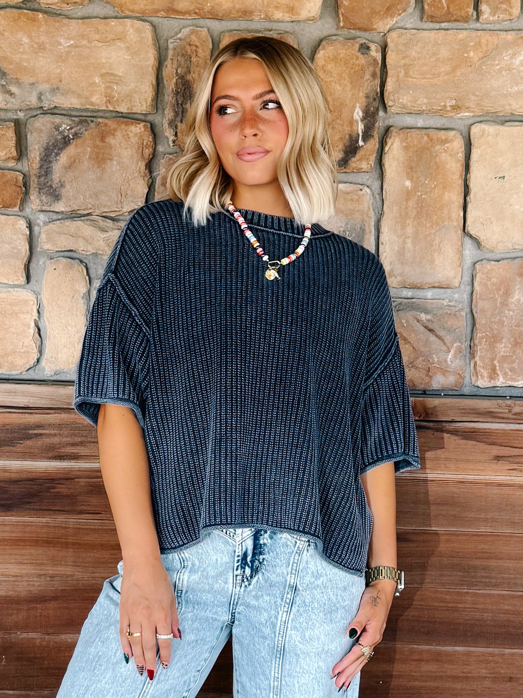 A woman with blonde hair stands in front of a stone wall wearing a dark navy blue ribbed knit top with wide short sleeves and light-wash blue jeans.