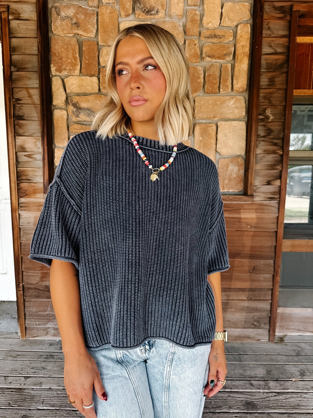 A woman with blonde hair wearing a Navy ribbed short-sleeve top and light blue jeans, standing on a wooden porch in front of a stone wall.