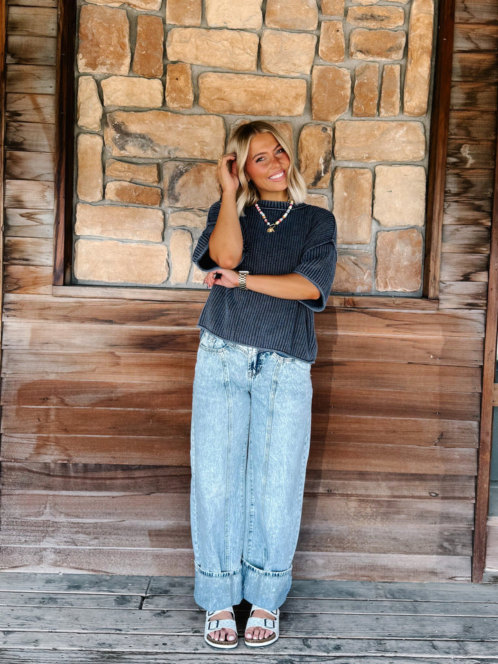 A woman stands outdoors against a rustic stone and wood wall, wearing a navy ribbed knit top, light-wash wide-leg jeans, and white slide sandals.