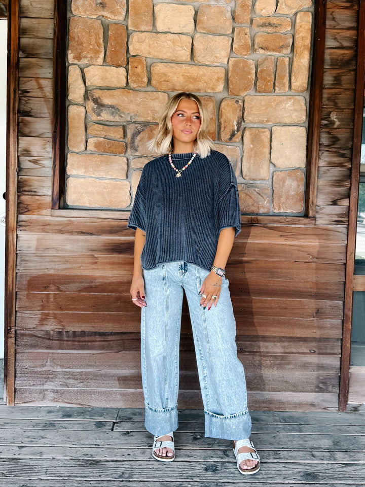A woman stands on a wooden deck in front of a rustic stone wall, wearing a dark navy blue ribbed short-sleeve top, light-wash wide-leg jeans, and white slide sandals.